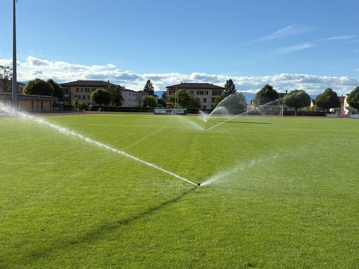 Terrain de football arrosé par des arroseurs à grande portée, tribune et village en arrière-plan
