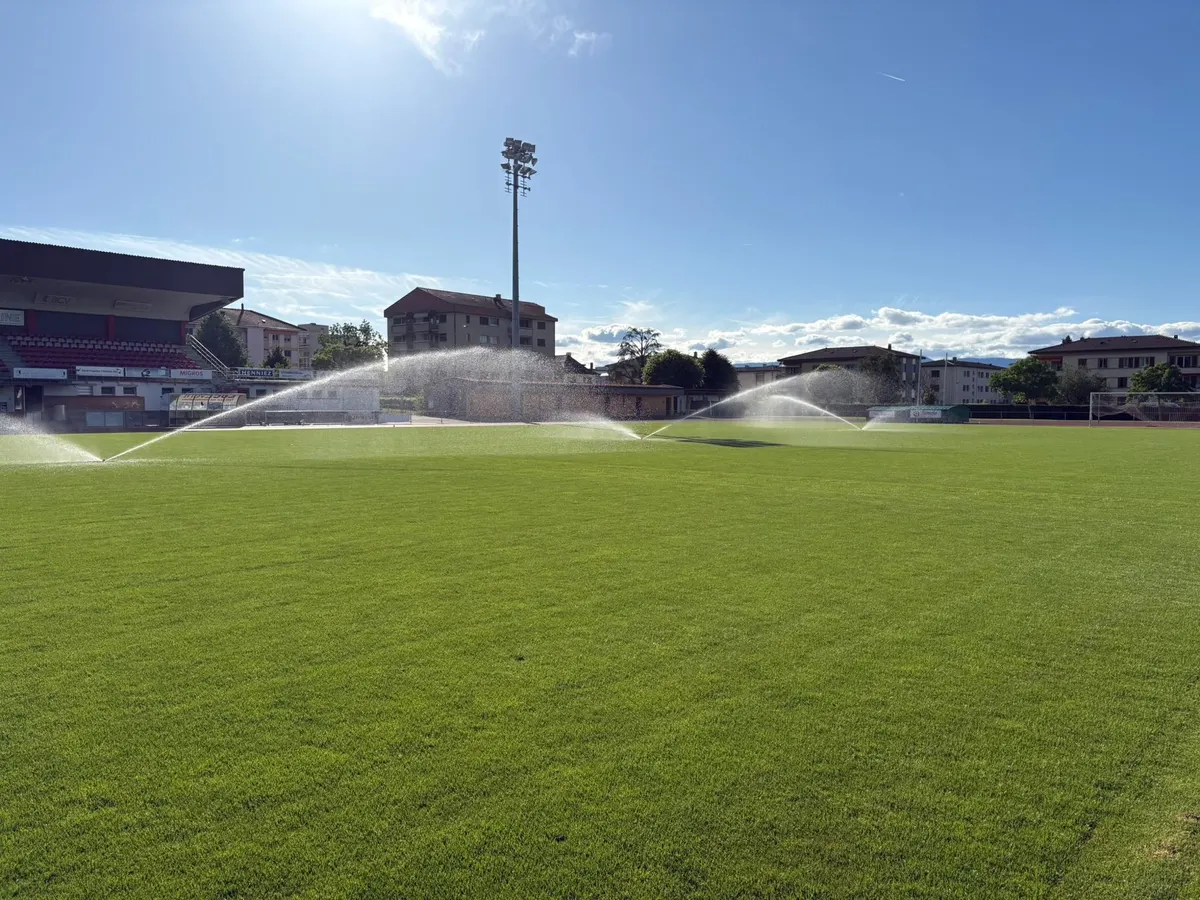 Stade de football avec tribune couverte, plusieurs arroseurs en fonctionnement sur la pelouse