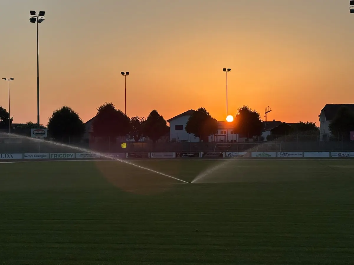 Arrosage nocturne d’un terrain de football au coucher du soleil, projecteurs en silhouette